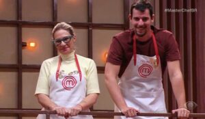 Dois participantes do MasterChef Brasil 2025, Daniela e Felipe B., aparecem lado a lado usando aventais brancos com o logo do programa. Daniela está à esquerda, com óculos de armação verde e blusa clara, enquanto Felipe está à direita, sorrindo, com camiseta vinho. Ambos apoiam as mãos no corrimão e parecem emocionados no estúdio iluminado.
