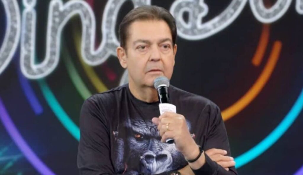 Foto de Fausto Silva, apresentador conhecido como Faustão, segurando um microfone durante gravação em estúdio, com expressão séria e camisa preta estampada com imagem de gorila. Fundo desfocado com luzes coloridas