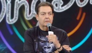 Foto de Fausto Silva, apresentador conhecido como Faustão, segurando um microfone durante gravação em estúdio, com expressão séria e camisa preta estampada com imagem de gorila. Fundo desfocado com luzes coloridas