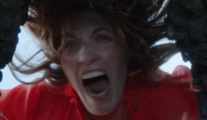 Florence Welch, vocalista do Florence + The Machine, em cena do teaser sombrio cavando um buraco, vestindo vestido vermelho, expressão intensa e gritando.