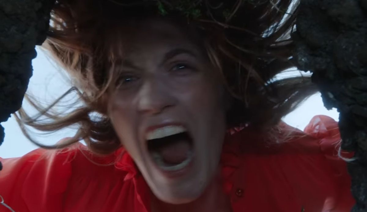 Florence Welch, vocalista do Florence + The Machine, em cena do teaser sombrio cavando um buraco, vestindo vestido vermelho, expressão intensa e gritando.