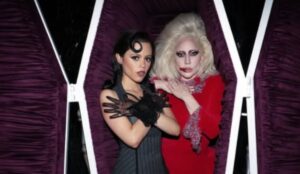 Jenna Ortega e Lady Gaga posam dentro de um caixão forrado de veludo roxo. Jenna está à esquerda, vestindo um look preto com luvas de tule e cabelo ondulado, fazendo o gesto de braços cruzados. Lady Gaga está à direita, com cabelo loiro volumoso, maquiagem dramática e vestido vermelho com detalhes brilhantes, também com os braços cruzados no peito. As duas encaram a câmera com expressão séria.