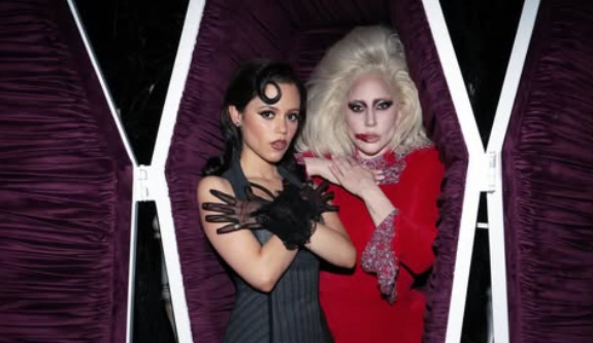 Jenna Ortega e Lady Gaga posam dentro de um caixão forrado de veludo roxo. Jenna está à esquerda, vestindo um look preto com luvas de tule e cabelo ondulado, fazendo o gesto de braços cruzados. Lady Gaga está à direita, com cabelo loiro volumoso, maquiagem dramática e vestido vermelho com detalhes brilhantes, também com os braços cruzados no peito. As duas encaram a câmera com expressão séria.