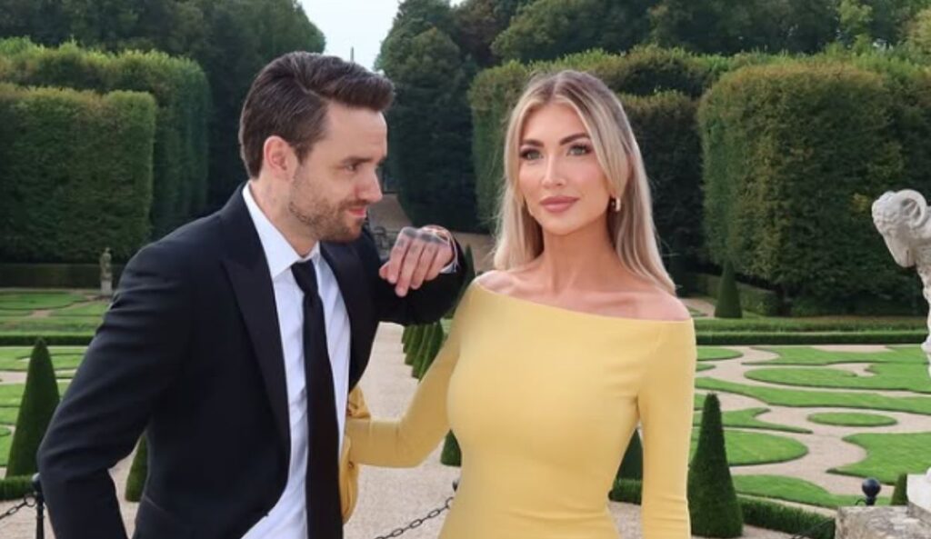 Liam Payne de terno preto e Kate Cassidy com vestido amarelo posam juntos em um jardim elegante com arbustos aparados e esculturas ao fundo.