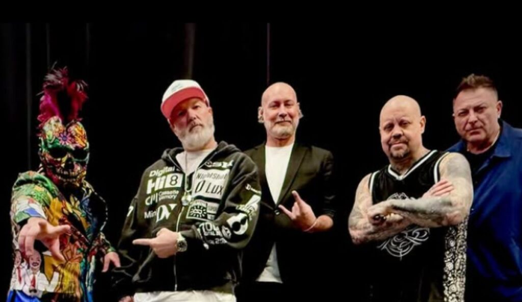 Integrantes do Limp Bizkit posam para foto promocional. Fred Durst está no centro, com barba grisalha, boné branco e casaco preto com estampas brancas. À esquerda, Wes Borland usa uma máscara colorida com moicano e roupa estampada. John Otto, Sam Rivers e Dj Lethal aparecem ao fundo, em trajes casuais ou esportivos, em frente a um fundo preto