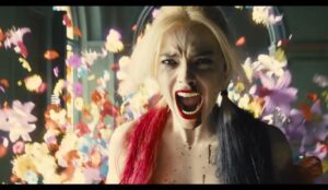 Margot Robbie como Arlequina, gritando com expressão de raiva e desespero, com flores coloridas voando ao fundo, em cena cheia de energia e caos visual.