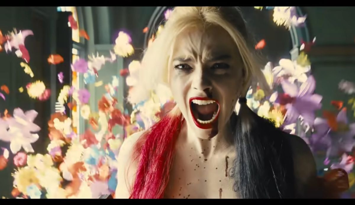 Margot Robbie como Arlequina, gritando com expressão de raiva e desespero, com flores coloridas voando ao fundo, em cena cheia de energia e caos visual.