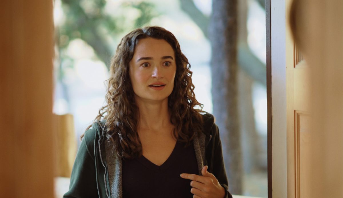 Kelley Mack em cena de produção, com expressão surpresa e vestida de forma casual, em ambiente iluminado por luz natural.
