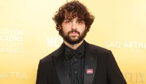 Noah Centineo em evento do SAG Awards, vestindo terno preto com gravata borboleta e fundo amarelo com logotipos da Netflix e SAG-AFTRA.