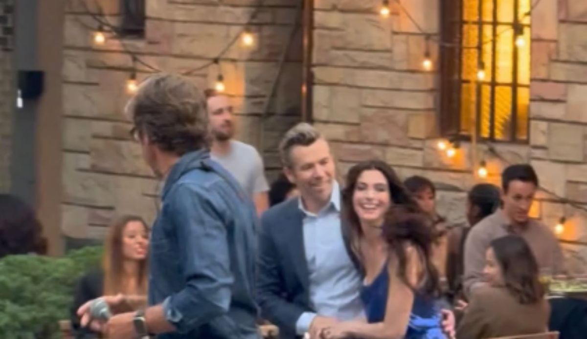 Anne Hathaway e Patrick Brammall gravam cena romântica de ‘O Diabo Veste Prada 2’ em set decorado com luzes, em Nova York. A atriz aparece sorridente, de vestido azul cerúleo, ao lado do ator australiano, sugerindo o clima leve entre os personagens