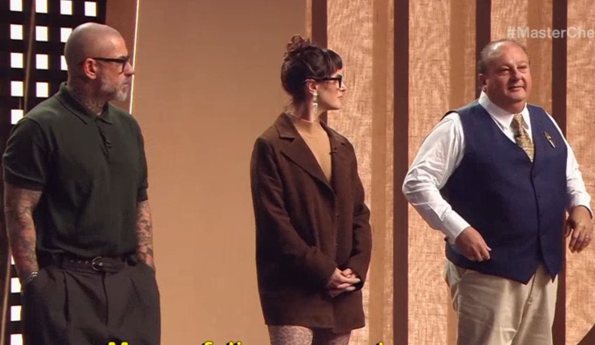 Três jurados do MasterChef Brasil 2026 estão lado a lado no estúdio: à esquerda, Henrique Fogaça usa barba, óculos e camisa polo preta; ao centro, Helan Rizzo aparece de cabelo preso com franja, óculos e blazer marrom; à direita, Erick Jacquin está sorridente usando colete azul, camisa branca e gravata estampada