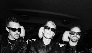 Integrantes da banda Rüfüs Du Sol sentados no banco traseiro de um carro, usando óculos escuros e jaquetas de couro, em uma foto em preto e branco.
