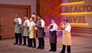 Seis participantes do reality Chef de Alto Nível estão lado a lado no estúdio do programa, usando aventais coloridos e aplaudindo durante a semifinal.