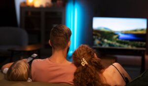 Família assistindo TV em uma sala aconchegante à noite; duas pessoas adultas e uma criança estão sentadas no sofá, vistas de costas, com a televisão exibindo uma paisagem colorida ao fundo.