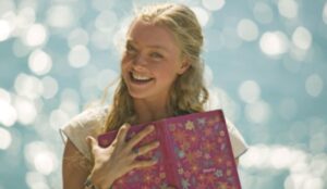 Amanda Seyfried como Sophie em cena de “Mamma Mia!”, sorrindo e segurando um diário colorido com o mar ao fundo