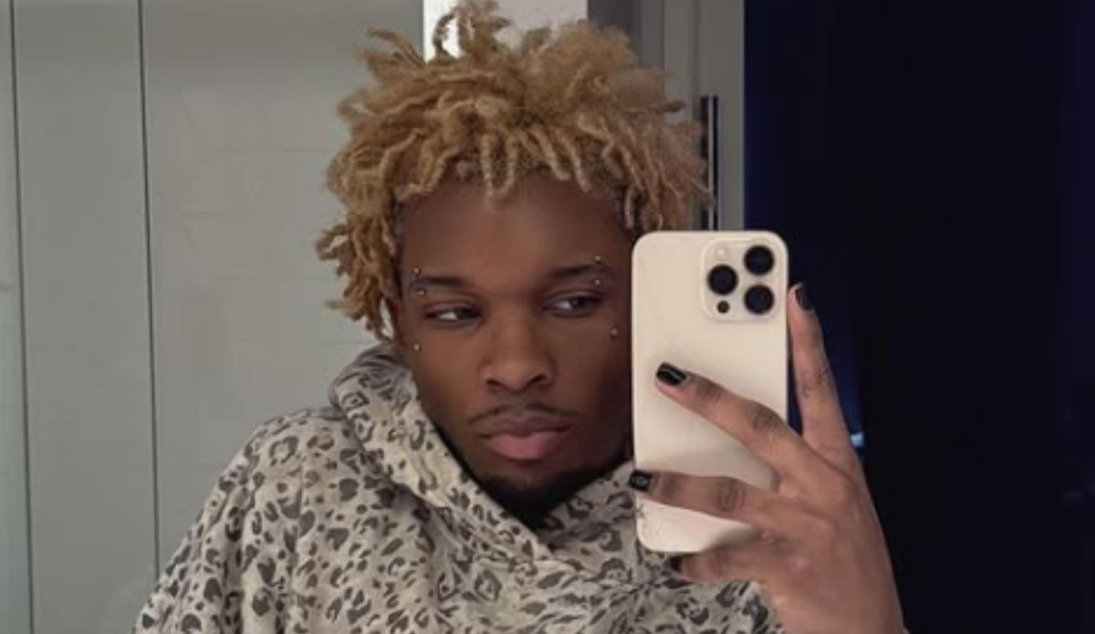 D4vd com cabelo loiro descolorido e estilo dread curto tira selfie em frente ao espelho usando moletom estampado de oncinha cinza. Ele segura um celular iPhone branco com capa, tem unhas pintadas de preto e piercings nas sobrancelhas e próximo aos olhos.