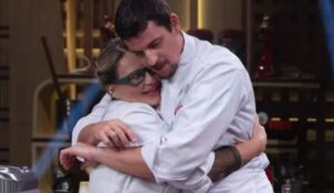 Daniela e Felipe B., os dois finalistas do MasterChef Brasil 2025 se abraçam na cozinha do programa, vestindo dólmãs brancos; ao fundo, utensílios e bancadas do cenário.