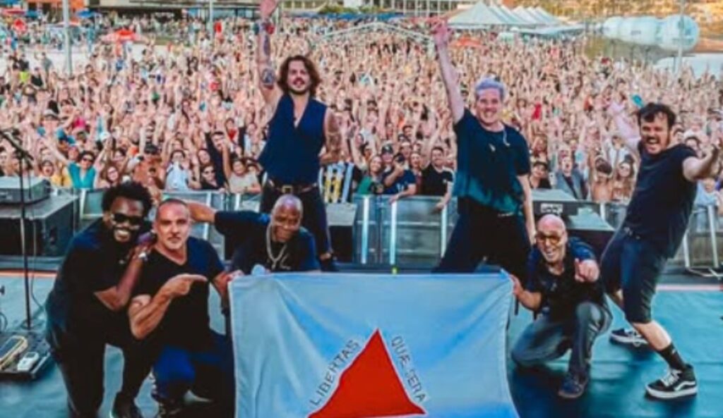 Banda Jota Quest posa no palco segurando a bandeira de Minas Gerais diante de uma multidão de fãs em show lotado, durante apresentação em festival de música; banda se apresenta no The Town 2025 nesta sexta-feira (12)