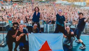 Banda Jota Quest posa no palco segurando a bandeira de Minas Gerais diante de uma multidão de fãs em show lotado, durante apresentação em festival de música; banda se apresenta no The Town 2025 nesta sexta-feira (12)