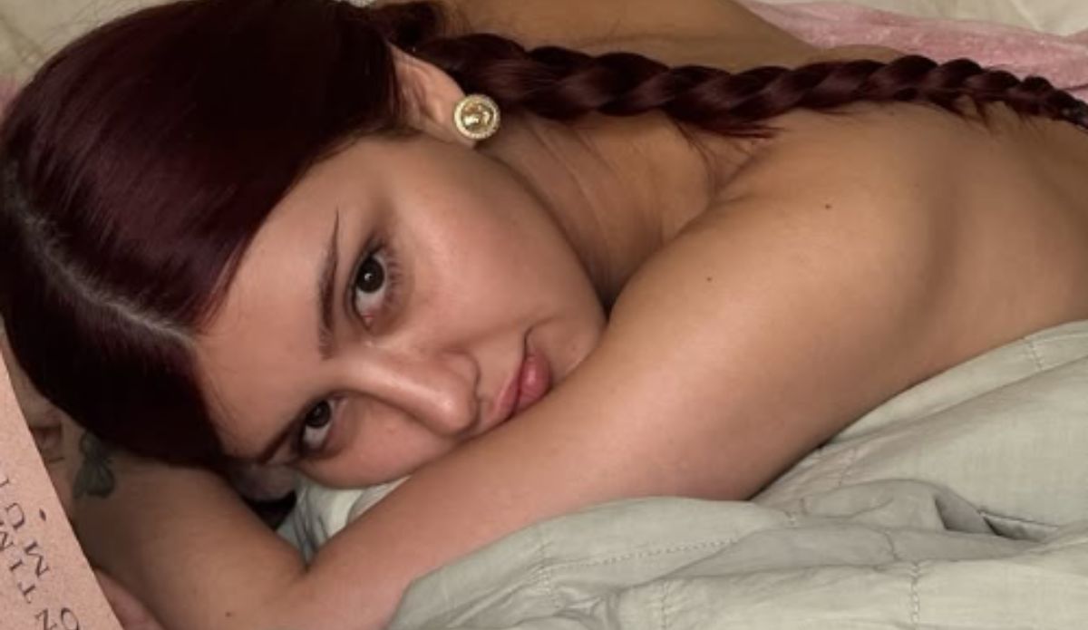 Sofia deitada de lado em uma cama, com tranças e brincos dourados, olha diretamente para a câmera em um retrato íntimo e próximo.