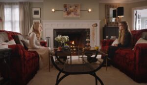 Amanda Seyfried e Sydney Sweeney em cena do filme “A Empregada”, sentadas em sofás vermelhos frente a uma lareira acesa.