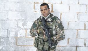 Cena promocional de Arcanjo Renegado mostra o personagem Mikhael em uniforme militar, segurando uma arma e usando colete tático diante de uma parede de concreto.