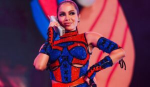 Anitta se apresenta em palco colorido, segurando um telefone antigo como parte da performance. Ela usa um figurino brilhante nas cores azul e vermelho, com luvas e top detalhados, durante show no Carnaval Na Cidade.