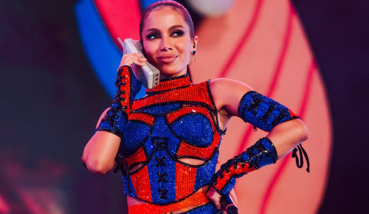 Anitta se apresenta em palco colorido, segurando um telefone antigo como parte da performance. Ela usa um figurino brilhante nas cores azul e vermelho, com luvas e top detalhados, durante show no Carnaval Na Cidade.