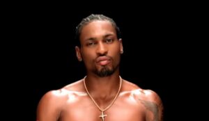 Cantor americano D’Angelo aparece em plano fechado, sem camisa e usando um colar com crucifixo, sobre fundo preto, em uma cena clássica de um de seus videoclipes.