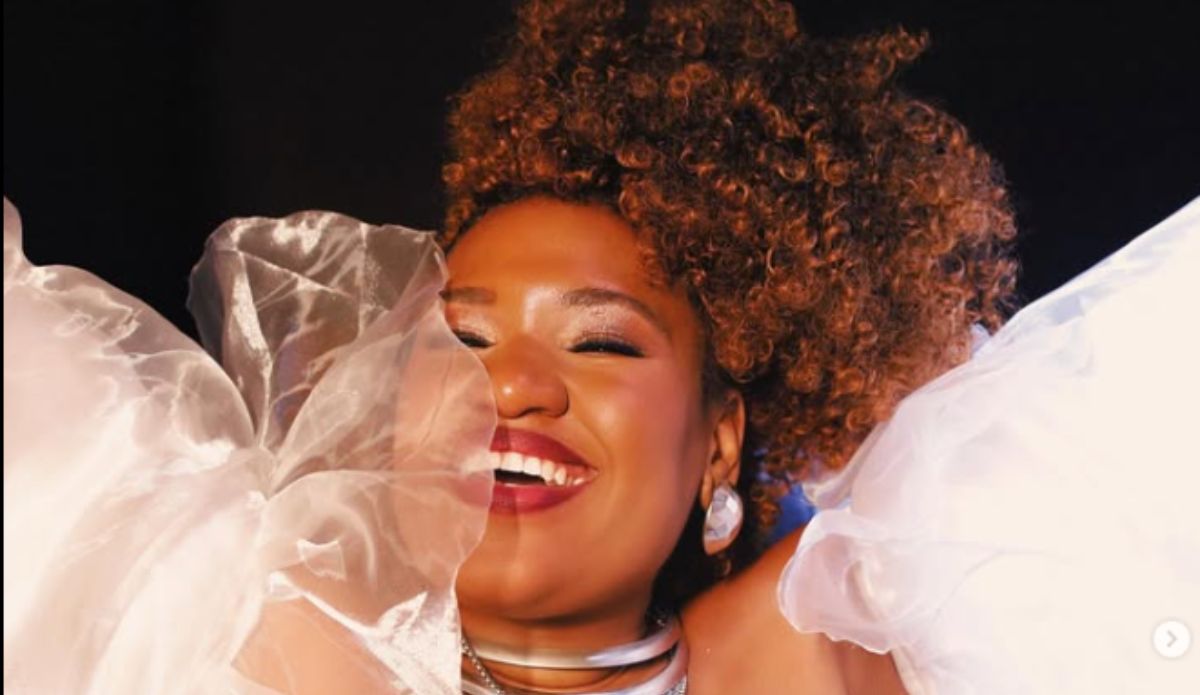 DJ Tamy sorri em retrato vibrante, usando look branco volumoso e maquiagem iluminada. A imagem transmite alegria, força e estilo — características marcantes da artista carioca.