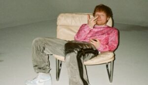 Cantor britânico Ed Sheeran sentado em uma poltrona bege, vestindo moletom rosa e calça esportiva cinza, em um ambiente minimalista e iluminado suavemente.