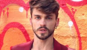O ator Felipe Selau posa em frente a uma parede colorida com grafite vermelho e laranja, vestindo um blazer vinho e com expressão séria.