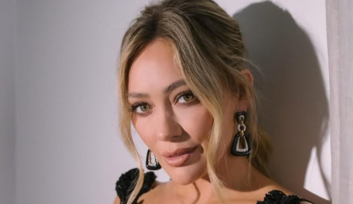 Hilary Duff posa com vestido preto e brincos grandes, olhando para a câmera com expressão suave; ao fundo, uma parede clara iluminada pela luz natural