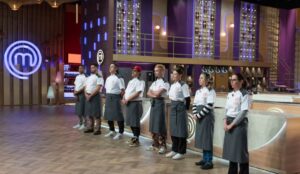 Oito participantes do MasterChef Confeitaria 2025 estão alinhados em fila no estúdio do programa, usando aventais cinza e camisetas brancas com o logotipo do reality. O cenário tem luzes roxas e douradas, e o símbolo do MasterChef iluminado aparece ao fundo.
