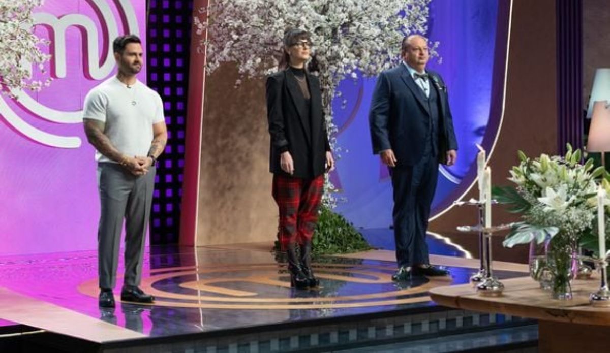Os jurados do MasterChef Confeitaria Brasil 2025 — Diego Lozano, Helena Rizzo e Erick Jacquin — posam no estúdio do programa durante o 7º episódio, com cenário decorado em tons de rosa, lilás e flores brancas.