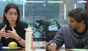 Michelle Barros e Shia Phoenix conversam sentados à mesa durante uma cena de “A Fazenda 17”, com clima sério e garrafa branca da Betano sobre a mesa.