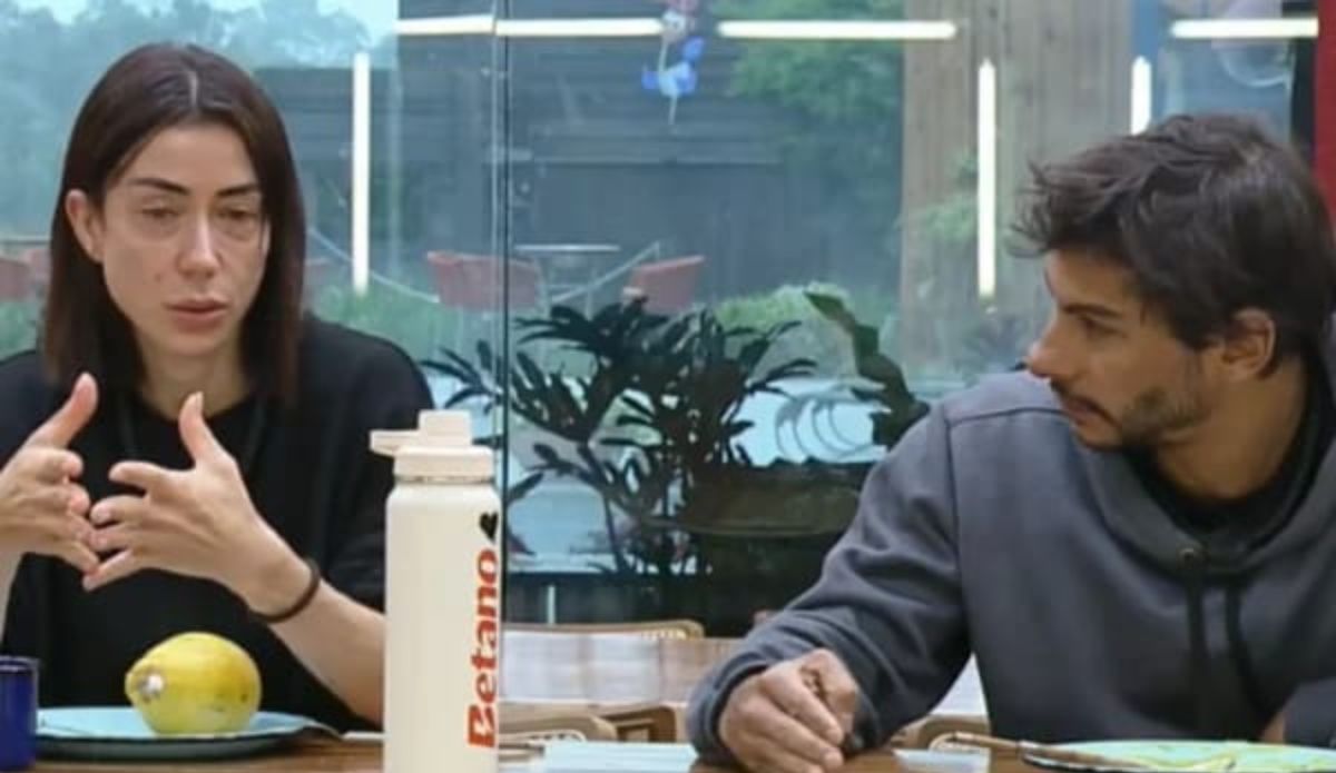 Michelle Barros e Shia Phoenix conversam sentados à mesa durante uma cena de “A Fazenda 17”, com clima sério e garrafa branca da Betano sobre a mesa.
