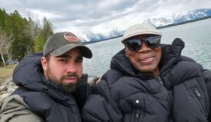 Milton Nascimento e o filho Augusto posam juntos à beira de um lago, cercados por montanhas nevadas. Ambos usam casacos grossos de frio; Milton sorri usando óculos escuros e boné claro, enquanto Augusto veste boné verde e olha para a câmera.