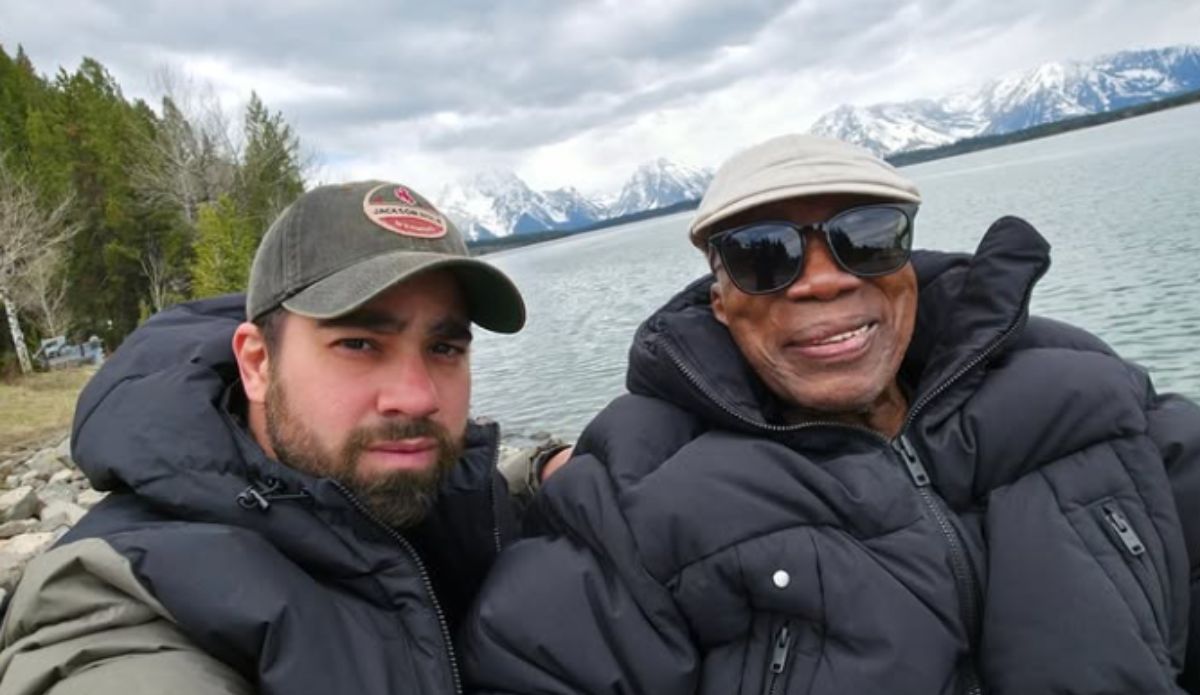 Milton Nascimento e o filho Augusto posam juntos à beira de um lago, cercados por montanhas nevadas. Ambos usam casacos grossos de frio; Milton sorri usando óculos escuros e boné claro, enquanto Augusto veste boné verde e olha para a câmera.