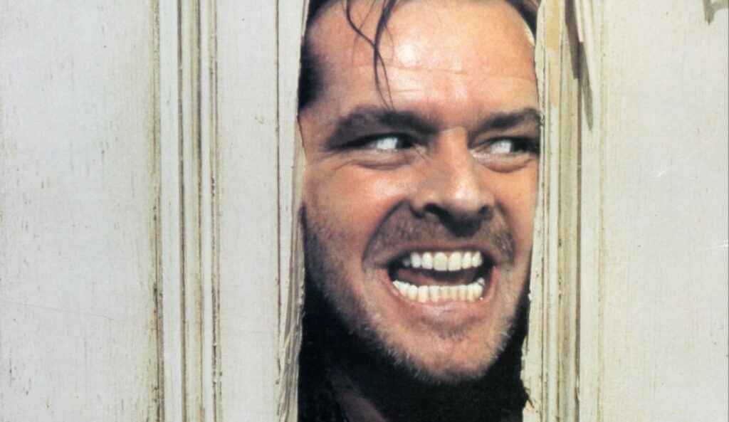 Cena icônica do filme O Iluminado mostra um homem com expressão insana espiando por uma porta quebrada, em um dos momentos mais marcantes do terror no cinema.