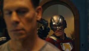 Cena de Pacificador mostra o herói usando seu capacete característico e apontando uma arma para outro homem, em um momento de tensão dentro de uma casa.