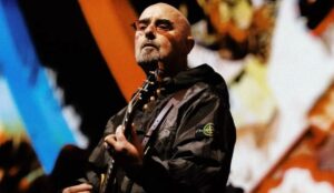 Paul Arthurs, guitarrista do Oasis, durante apresentação da turnê de reunião da banda. O músico veste jaqueta preta e segura sua guitarra sob luzes de palco