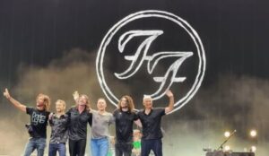 ntegrantes do Foo Fighters se despedem do público no palco, lado a lado e de mãos levantadas, com o logotipo “FF” da banda projetado em grande escala ao fundo.