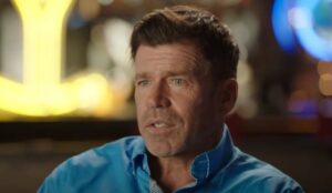 Taylor Sheridan, criador da série Yellowstone, aparece em entrevista usando uma camisa azul, com expressão séria e o fundo desfocado com luzes amareladas.