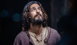 Cena da série “The Chosen” mostra o ator Jonathan Roumie caracterizado como Jesus, com barba e cabelos longos, olhando para cima em expressão contemplativa, usando túnica bege e manto roxo.