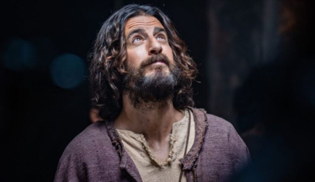 Cena da série “The Chosen” mostra o ator Jonathan Roumie caracterizado como Jesus, com barba e cabelos longos, olhando para cima em expressão contemplativa, usando túnica bege e manto roxo.