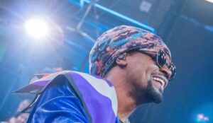 Toni Garrido, vocalista do Cidade Negra, sorridente no palco, usando óculos escuros, lenço colorido na cabeça e roupa azul brilhante, sob luz intensa no estúdio do Altas Horas.