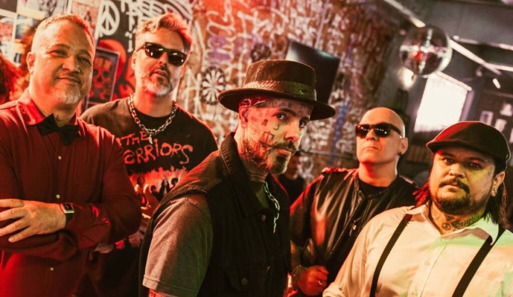 Integrantes do Detonautas posam em ambiente com estética underground, diante de uma parede repleta de grafites; ao centro, Tico Santa Cruz aparece usando chapéu preto e colete, enquanto os outros membros da banda surgem ao redor, iluminados por luzes avermelhadas.