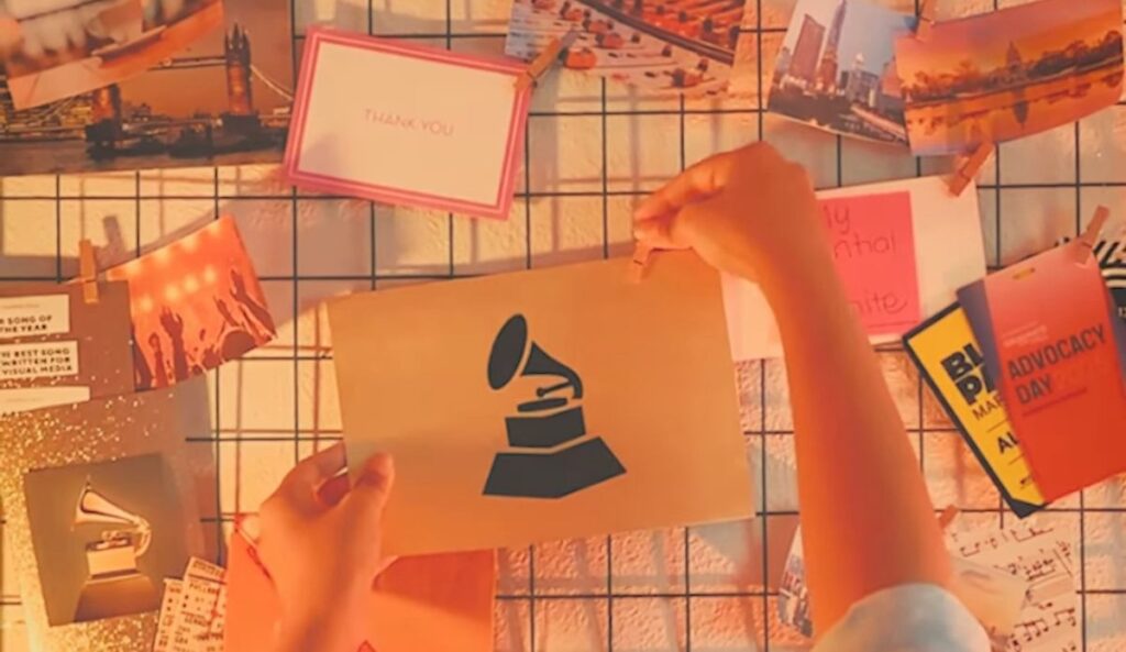 Mãos pendurando um cartão com o logotipo do Grammy em um painel de grade metálica, cercado por fotos, bilhetes e cartões coloridos sob uma luz quente e alaranjada.