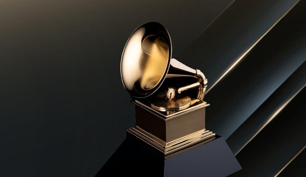 Imagem do troféu dourado do Grammy, em formato de gramofone, sobre uma base preta, com fundo escuro e detalhes metálicos iluminados.